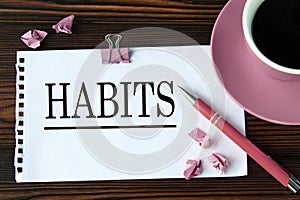 HABITS - word on a white sheet on a wooden brown background with a cup of coffee and a pen