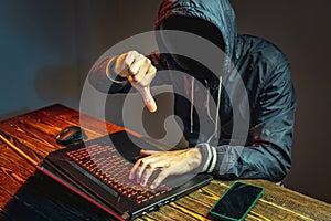 A hacker anonymous in a hood is typing on a laptop keyboard in a dark room. Cybercrime fraud and identity theft