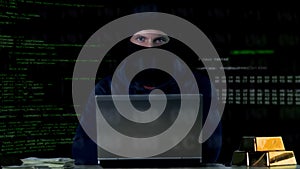 Hacker in balaclava committing cyber crime on laptop, numbers codes background