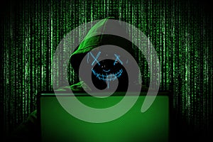 Hacker with glowing mask behind notebook laptop in front of green source binary code background internet cyber hack attack concept