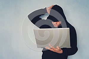 Hacker with his computer thinking on blue background. hacker in a black hood with a gray modern ultrabook, identity theft.