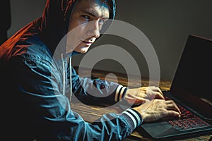 Hacker in a hood with a phone is typing on a laptop keyboard in a dark room. Concept of cyber warfare and Dos attacks