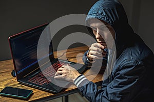 Hacker in a hood with a phone is typing on a laptop keyboard in a dark room. Cybercrime fraud and identity theft