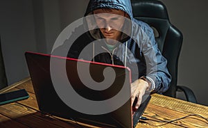 Hacker in a hood with a phone is typing on a laptop keyboard in a dark room. Cybercrime fraud and identity theft