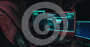 Hacker in a hood sits in front of computer screens and hacks databases