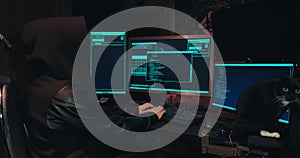 Hacker in a hood sits in front of computer screens and hacks databases