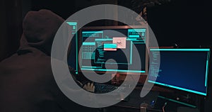 Hacker in a hood sits in front of computer screens and hacks databases
