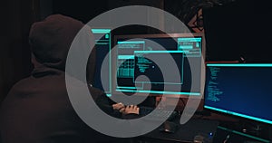 Hacker in a hood sits in front of computer screens and hacks databases