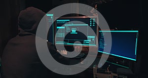 Hacker in a hood sits in front of computer screens and hacks databases