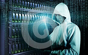 Hacker in Hoodie Standing in the Middle of Data Center full of Rack Servers and Hacking it with His Laptop.