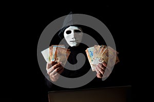 hacker man dressed in black with white mask and black hood holding euro banknotes in his hands