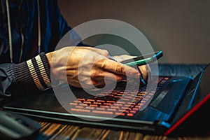 A hacker with a phone is typing with his hands on a laptop keyboard in a dark room. Cybercrime fraud and identity theft