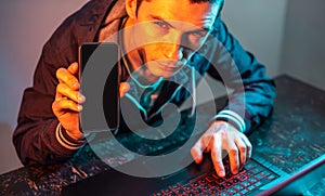 Hacker with phone is typing on a laptop keyboard in a dark room under a neon light. Cybercrime fraud and identity theft