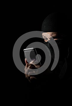 Hacker Using Smart Phone. Young adult girl in black clothes with hidden face looks at smartphone screen on black background with c