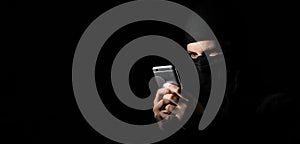 Hacker Using Smart Phone. Young adult girl in black clothes with hidden face looks at smartphone screen on black background with c