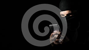 Hacker Using Smart Phone. Young adult girl in black clothes with hidden face looks at smartphone screen on black background with c