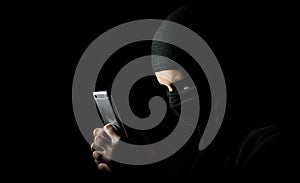 Hacker Using Smart Phone. Young adult girl in black clothes with hidden face looks at smartphone screen on black background with c