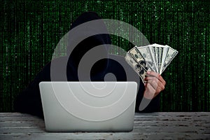Hacker wearing black hooded with computer hacking data and showing money on hand with green binary code background