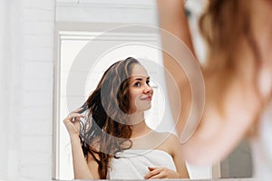 Hair and body care. Woman touching wet hair and smiling while looking in the mirror. Portrait of girl  in bathroom applying condit