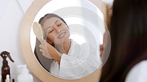 Hair Care concept. Young cheerful asian woman brushing her hair, looking at mirror at bathroom and smiling