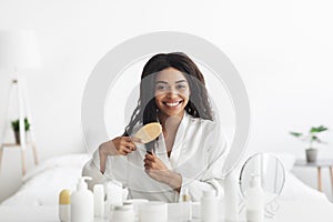 Daily hair care at home during covid-19 lockdown. Happy african american lady combing her curly hair in bedroom photo