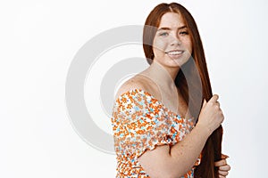 Hair care and women beauty. Young redhead woman touching her long red hair and smiling at camera, standing in dress over