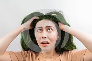 Hair coloring. A young Caucasian woman in a beige t-shirt, looking up at her long green hair in fright. White background