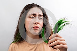 Hair coloring. A young Caucasian woman in a beige t-shirt looks distressed at the green tips of her long hair. White background