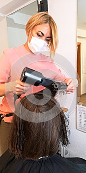 Hair drying with dryer in beauty salon.Blonde hairdresser in medical face mask,brunette client.Reopening,quarantine