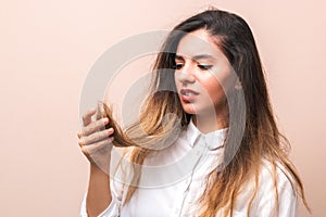 Hair problems. young woman in white shirt checking her britle, damaged, and split hairs against pink background