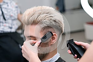 Haircut process of blond young man with hair trimmer in armchair in barbershop salon