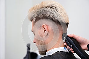 Haircut process of blond young man with hair trimmer in armchair in barbershop salon