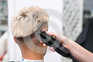 Haircut process of blond young man with hair trimmer in armchair in barbershop salon