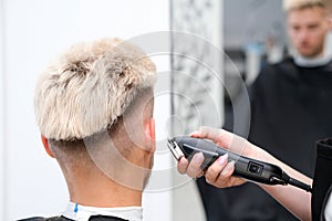 Haircut process of blond young man with hair trimmer in armchair in barbershop salon