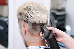 Haircut process of blond young man with hair trimmer in armchair in barbershop salon, barbershop concept