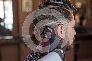 A hairdresser cuts a bearded young guy with a hair trimmer, combing the hair on his head. Work of the master in men`s haircut in