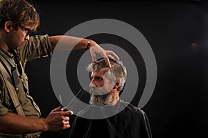 Hairdresser cuts senior citizen with a beard on a dark background