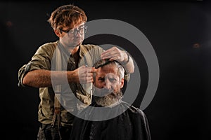 Hairdresser cuts senior citizen with a beard on a dark background