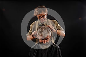 Hairdresser cuts senior citizen with a beard on a dark background