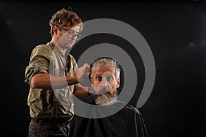 Hairdresser cuts senior citizen with a beard on a dark background