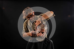 Hairdresser cuts senior citizen with a beard on a dark background