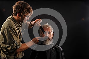 Hairdresser cuts senior citizen with a beard on a dark background