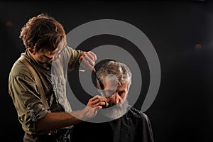 Hairdresser cuts senior citizen with a beard on a dark background