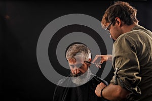 Hairdresser cuts senior citizen with a beard on a dark background