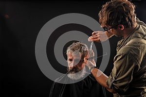 Hairdresser cuts senior citizen with a beard on a dark background