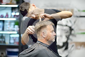 Hairdresser is cutting hair of handsome bearded mature man in salon. Stylist making hairstyle with electric shaver for person in
