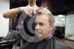 Hairdresser is cutting hair of handsome bearded mature man in salon. Stylist making hairstyle for person in barbershop. Services