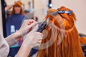 The hairdresser does hair extensions to a young, red-haired girl, in a beauty salon.