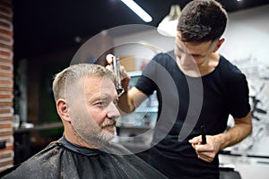 Hairdresser is doing eyebrow trimming using a razor for handsome bearded mature man in salon. Stylist making hairstyle for client