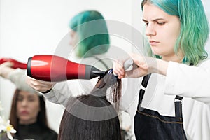 Hairdresser with green hair dries hair to client with red hairdryer, blue comb in beauty salon